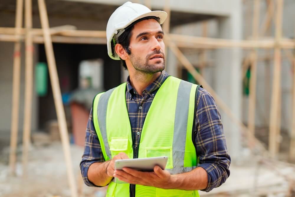 Construction worker in green vest holding tablet and looking upwards.