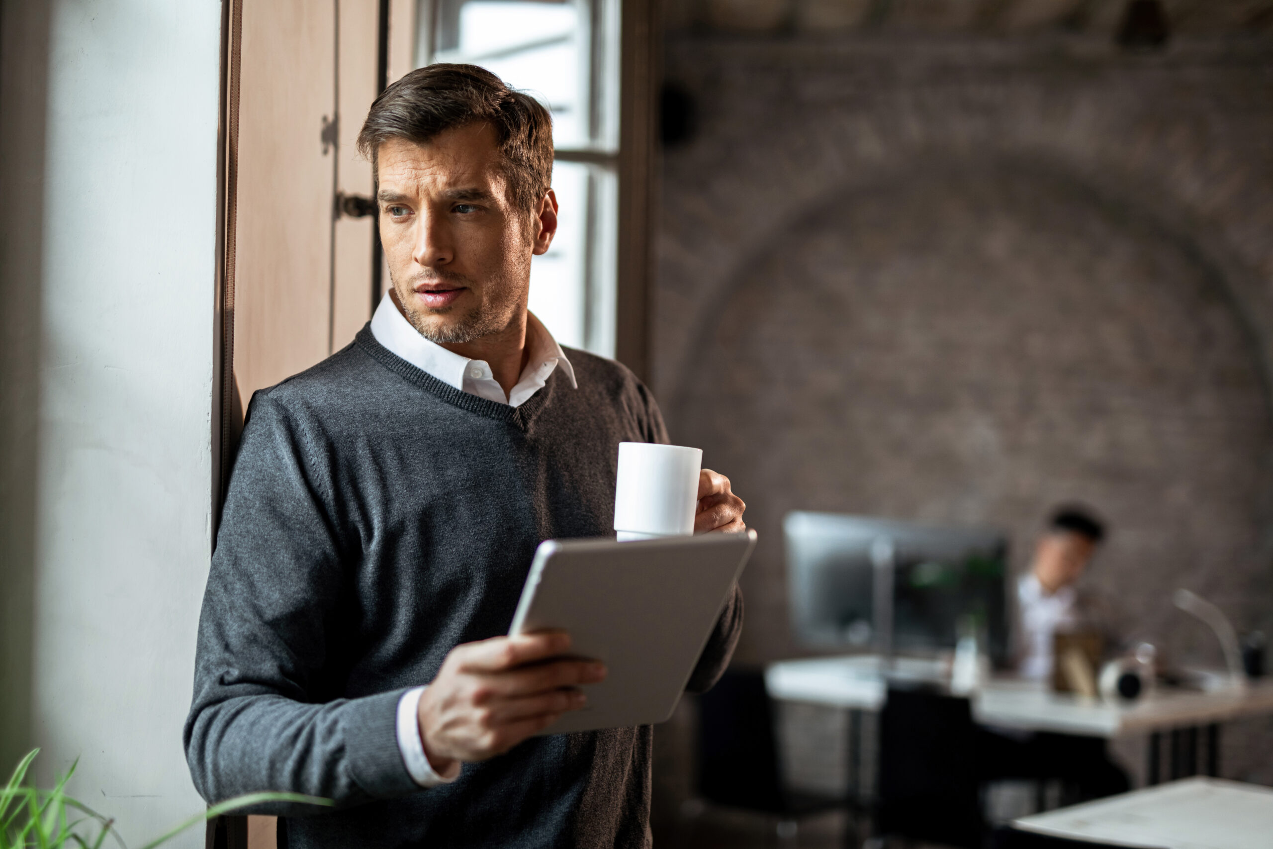 man holding tablet and coffee cup while looking out office window